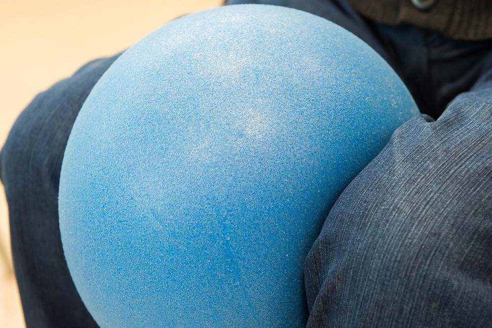 Blauer Gymnastikball ruht auf den Knien einer Person in blauer Jeans. Unschärfe im Hintergrund.